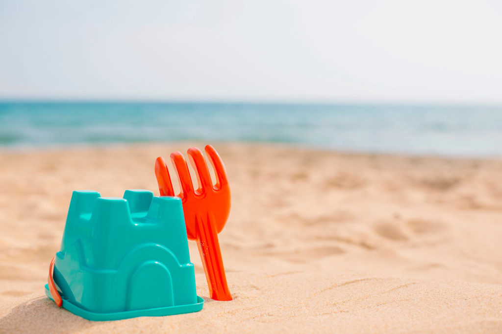 foto de una playa, con un cubo de plastico y un rastrillo clavados en la arena, de fondo se ve el mar
